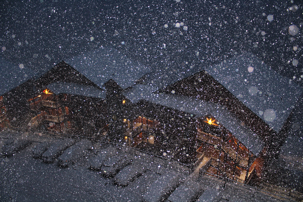 snow falling in front of wood buildings with snow covered roofs 