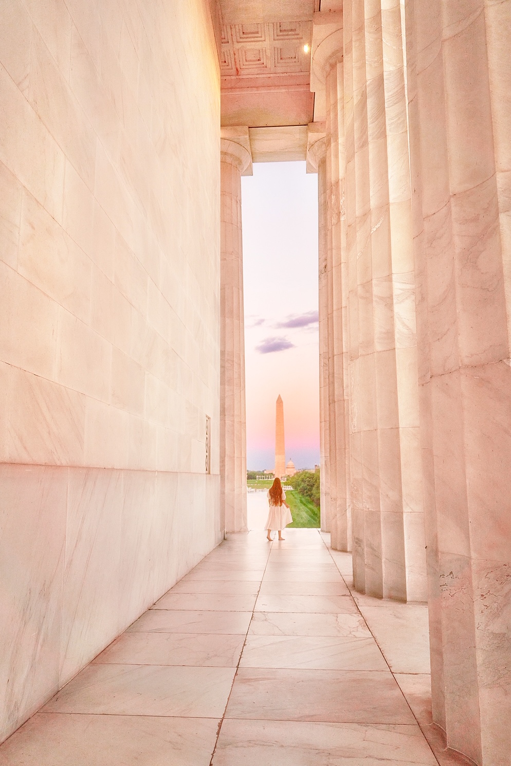 Looking down the corridor of the Lincoln Memorial. At the end of the corridor there is a woman wearing a white dress with long hair standing facing the Washington Monument in the distance. 