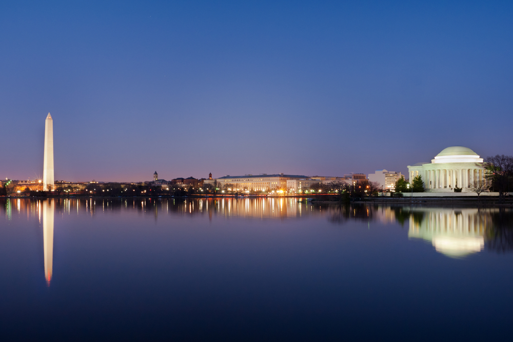 The Washington Monument, the Jefferson Memorial, and other buildings lit up at night. The view is of the monuments from across a large body of water. 