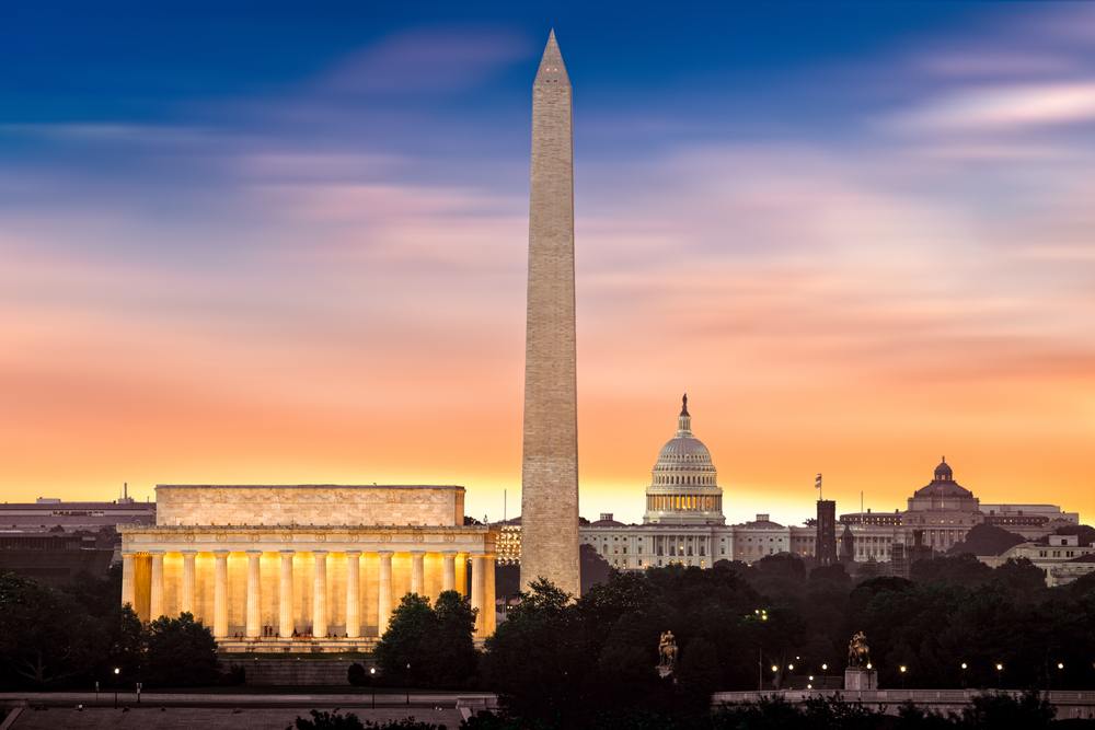 The monuments and memorials in the Washington DC skyline. It is twilight, so the buildings are lit up. The sky is blue, purple, pink, orange, and yellow. 