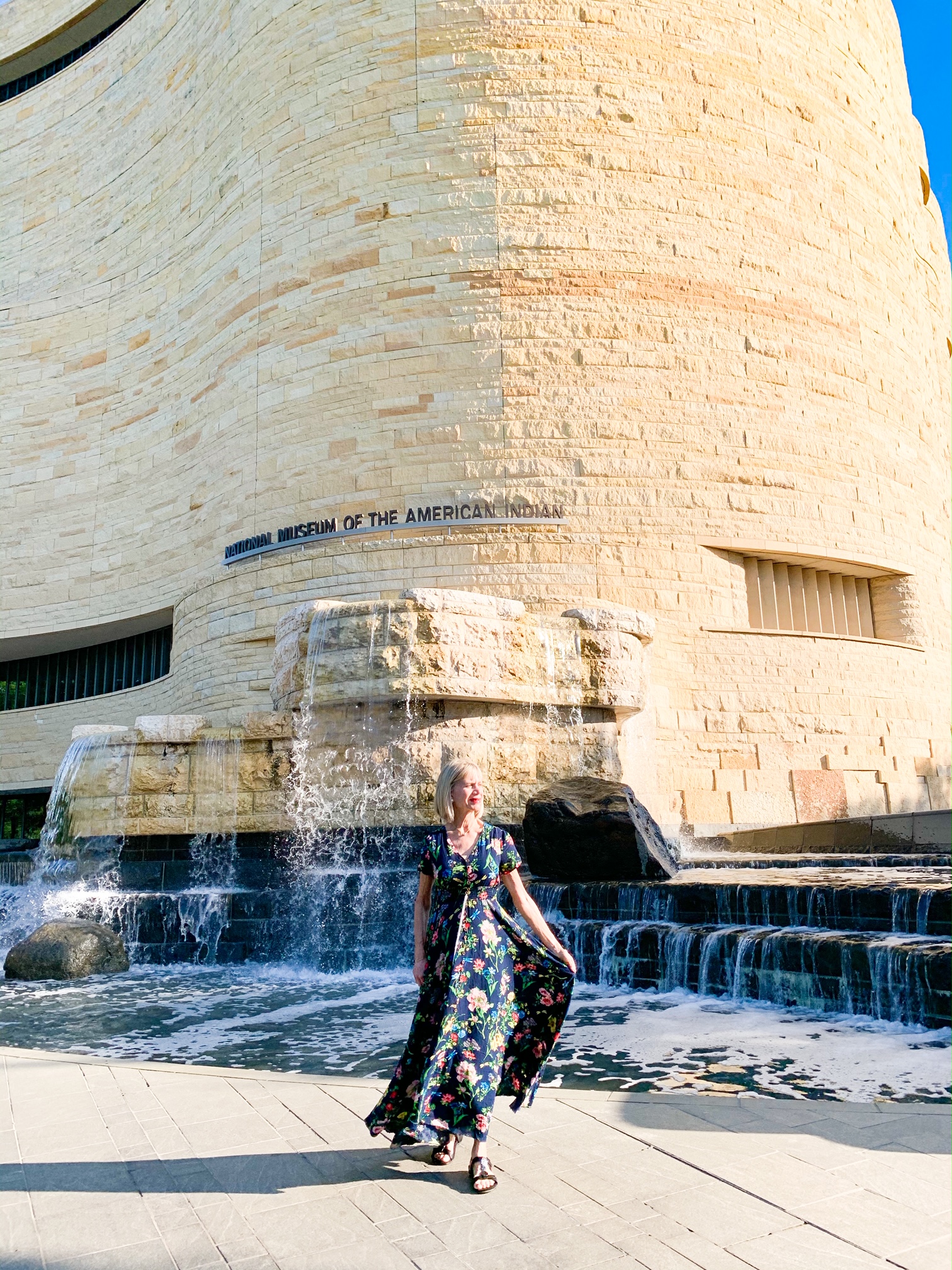 A woman in a dark floral dress standing in front of a fountain that looks like a waterfall. It is in front of and partially connected to the National Museum of the American Indian. The building is made of what looks like rough cut sandstone bricks. 