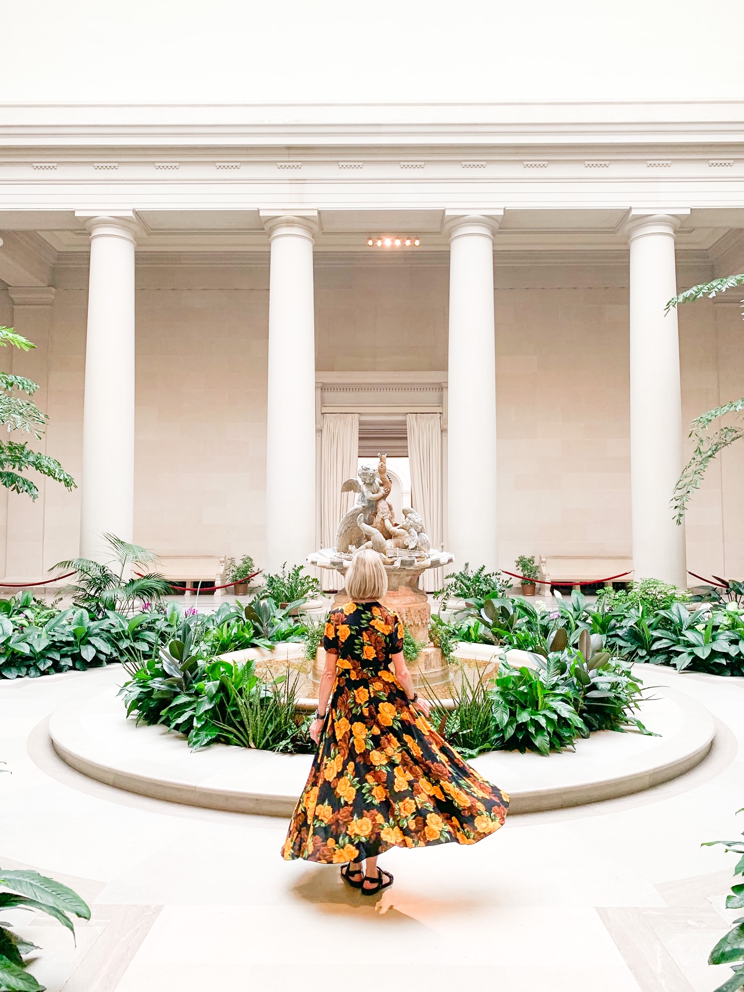 A woman in a dark floral dress looking away from the camera towards a fountain in the middle of a courtyard. The courtyard has lots of greenery and roman columns around a hallway that leads to other rooms. 