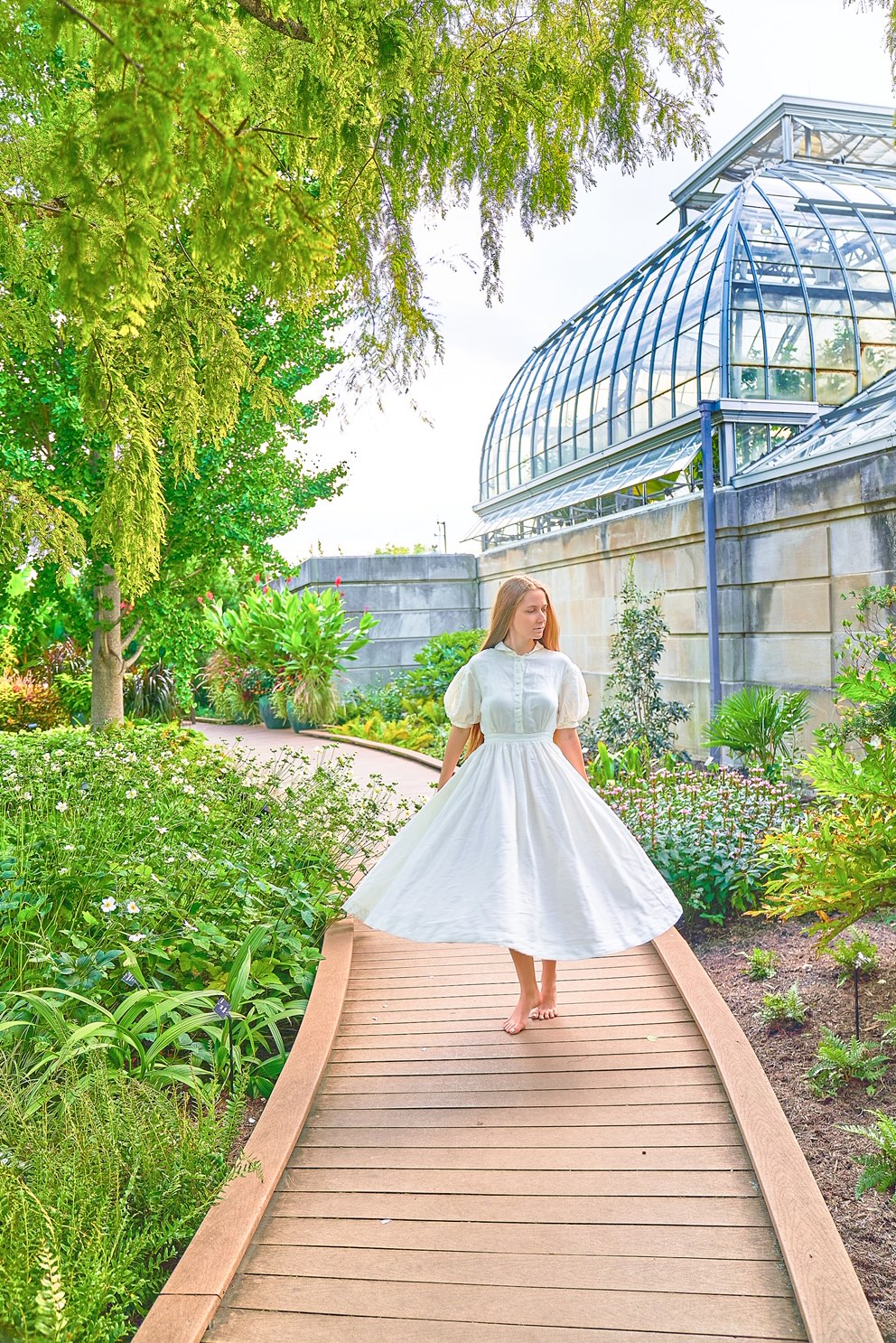 A woman in a white dress standing on a wooden walkway. She is surrounded by different plants and trees. You can see a conservatory building in the background. 