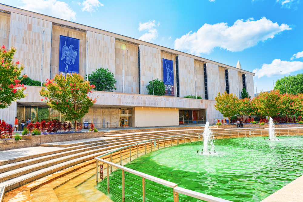 The front exterior of the National Museum of American History. It is a modern looking building with a wide courtyard and a water fountain in front of it. 