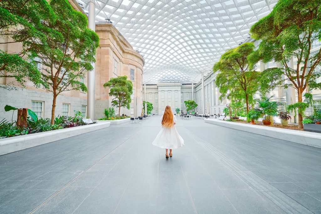 A woman in a white dress with long hair walking in the courtyard of the National Portrait Gallery, one of the best things to do in Washington DC. There are trees, shrubs, a wavy glass ceiling, and building facades.