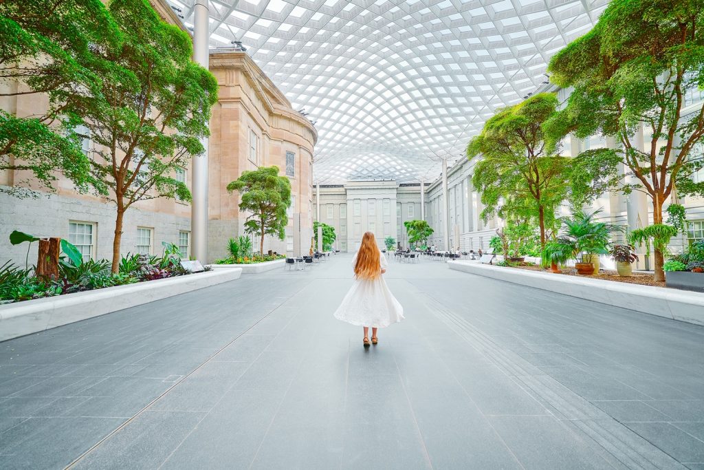 A woman in a white dress with long hair walking in the courtyard of the National Portrait Gallery, one of the best things to do in Washington DC. There are trees, shrubs, a wavy glass ceiling, and building facades. 