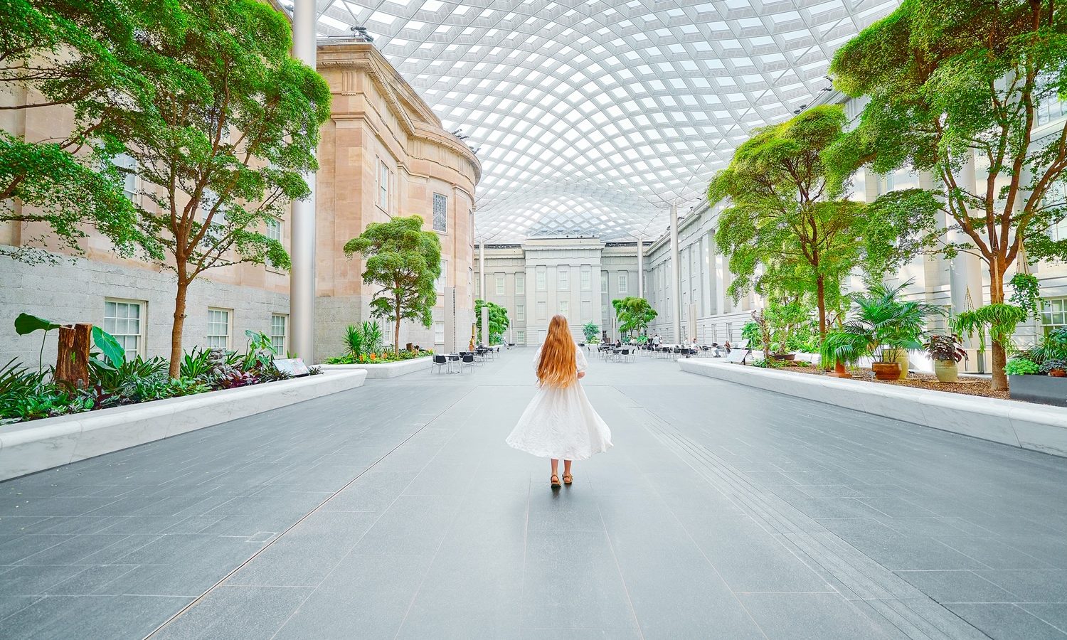 A woman in a white dress with long hair walking in the courtyard of the National Portrait Gallery, one of the best things to do in Washington DC. There are trees, shrubs, a wavy glass ceiling, and building facades.