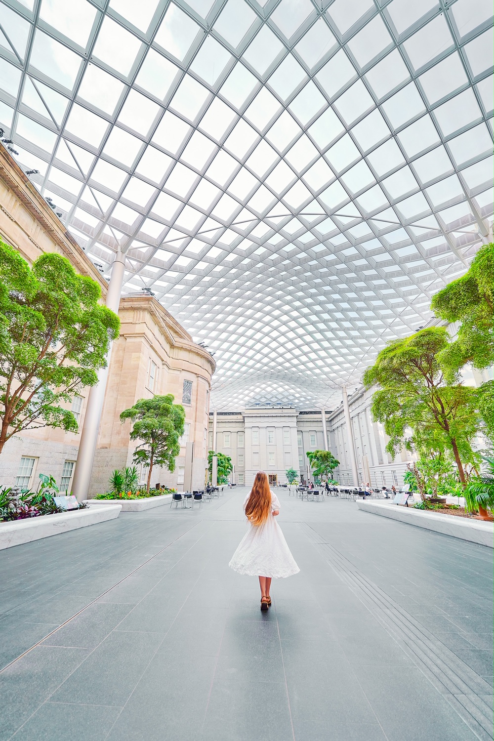 A woman in a white dress with long hair walking in the courtyard of the National  Portrait Gallery. There are tall trees, shrubs, bistro tables, building facades, and a wavy glass ceiling. 