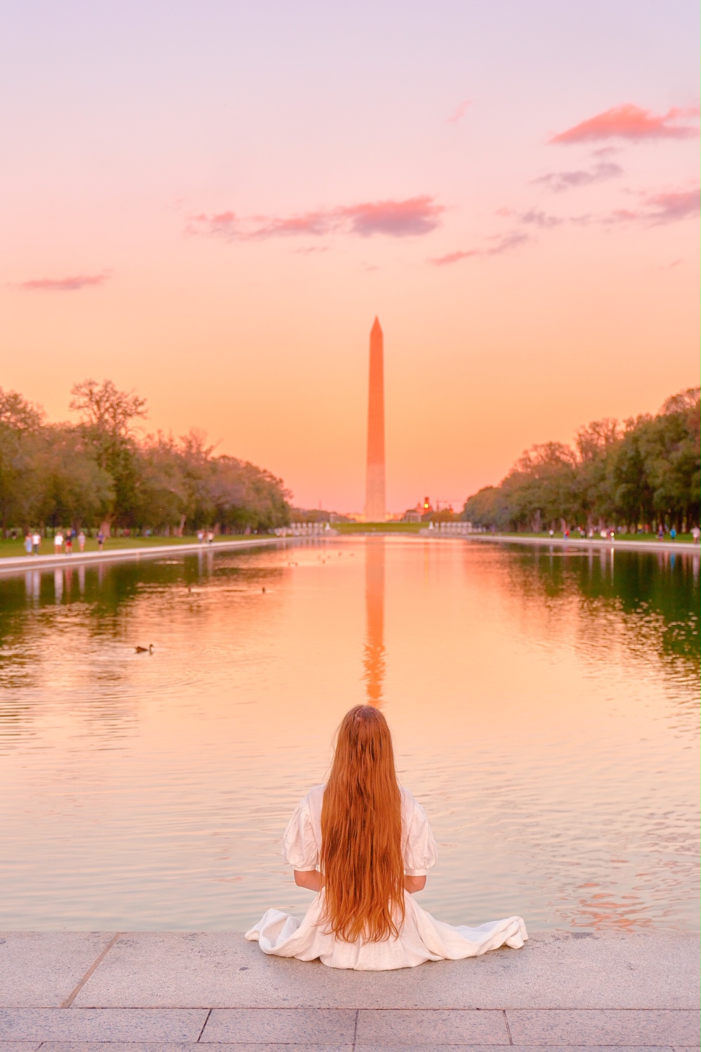 A woman in a white dress with long hair sitting in front of the reflecting pool in the National Mall. She is facing the Washington Monument and the sun setting. It's a must-stop place during any weekend in Washington DC. 