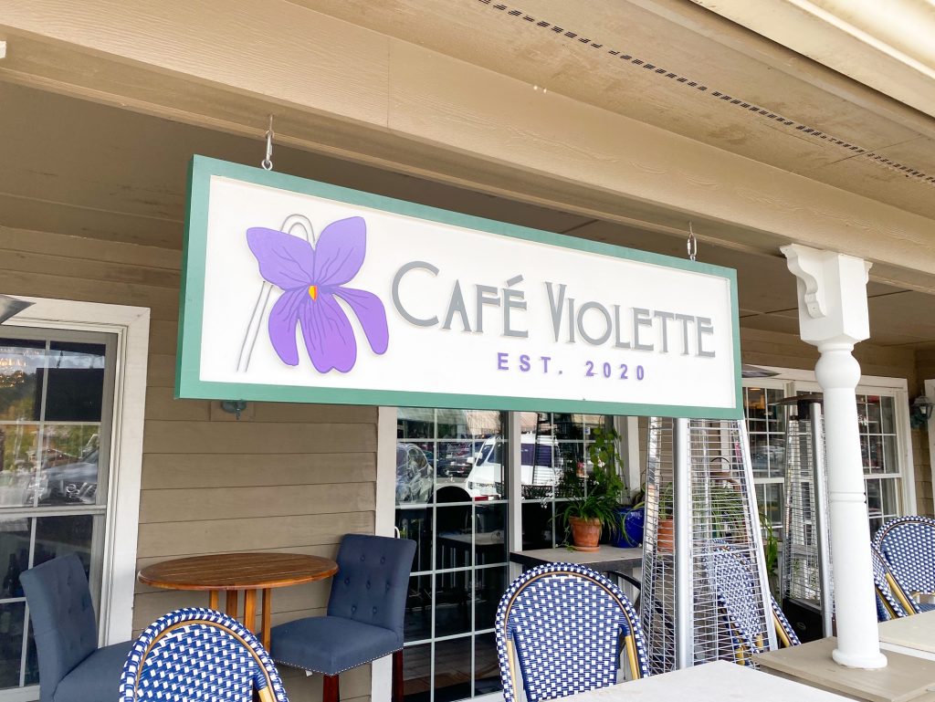 The sign in front of Cafe Violette with blue and white checkered chairs and tables