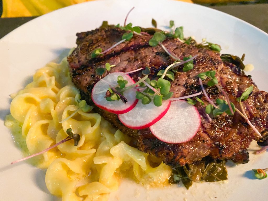 A grilled steak topped with radish and micro greens with noodles on a white plate served at one of the restaurants in Blowing Rock