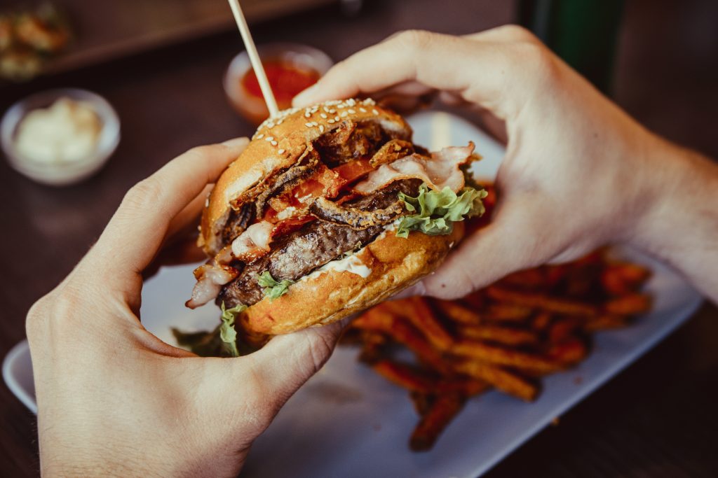 hands hold a burger with fries in the background