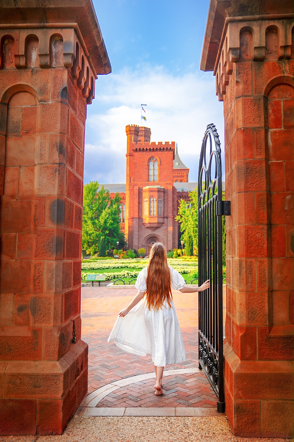 A woman in a white dress with long hair walking through the gate towards a garden. The garden has a brick pathway and there are yellow flowers. You can also see part of a large brick castle. Its one of the best things to do in Washington DC.