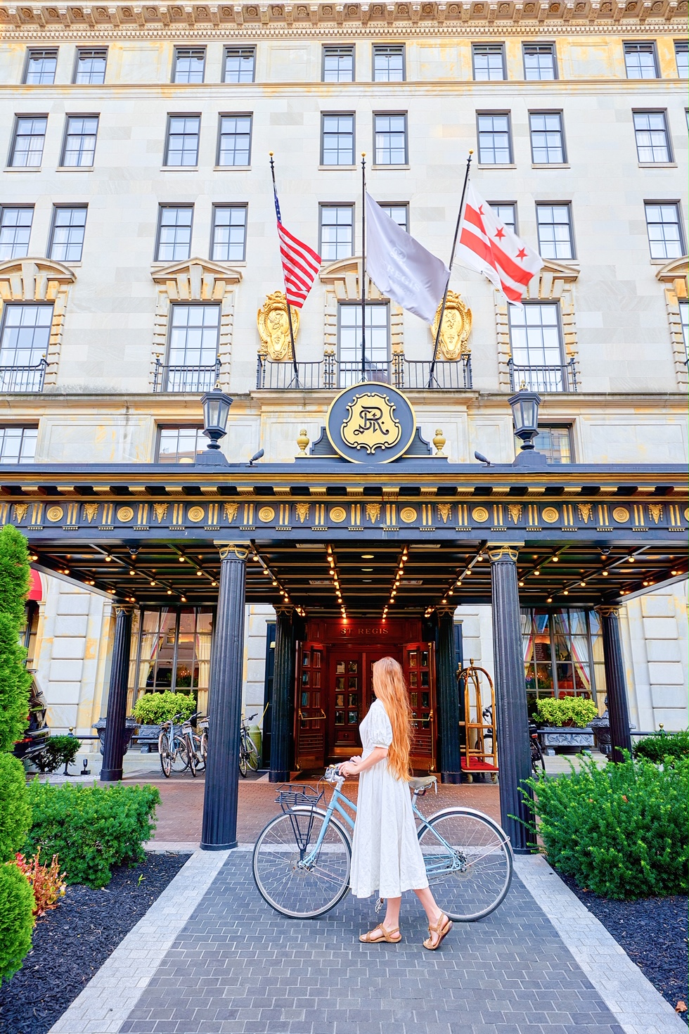 A woman in a white dress with long hair standing next to a bicycle. She is standing in front of the St. Regis Hotel in Washington DC. It is a ornately decorated and large hotel made of sandstone.