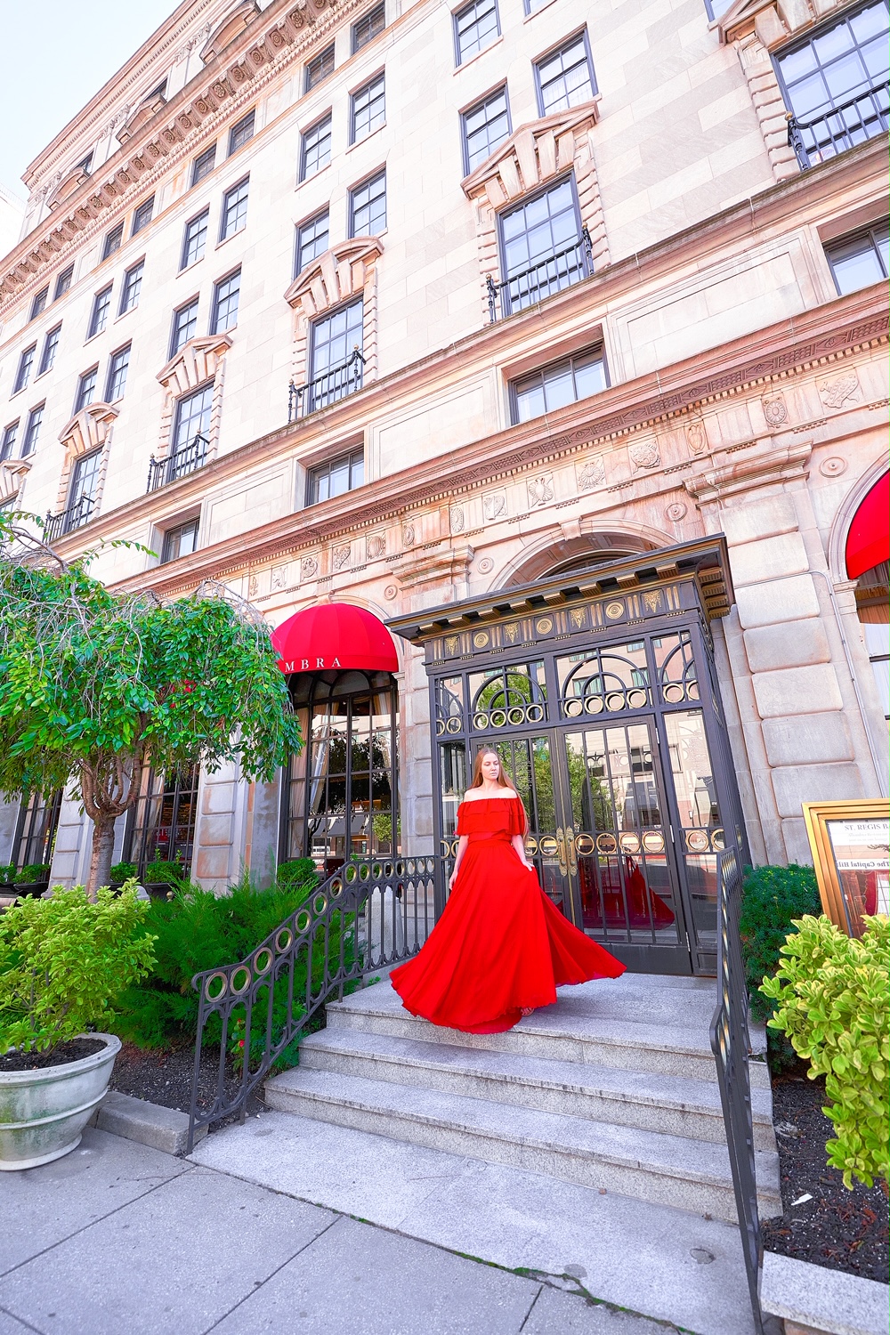 A woman in a long red dress standing on the steps of the St. Regis Hotel. It is an ornately decorated and historic hotel in DC. 