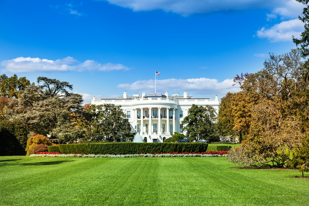 A view of the White House from across the White House Lawn. There is a large green lawn, lots of trees, shrubs, and some red flowers. 