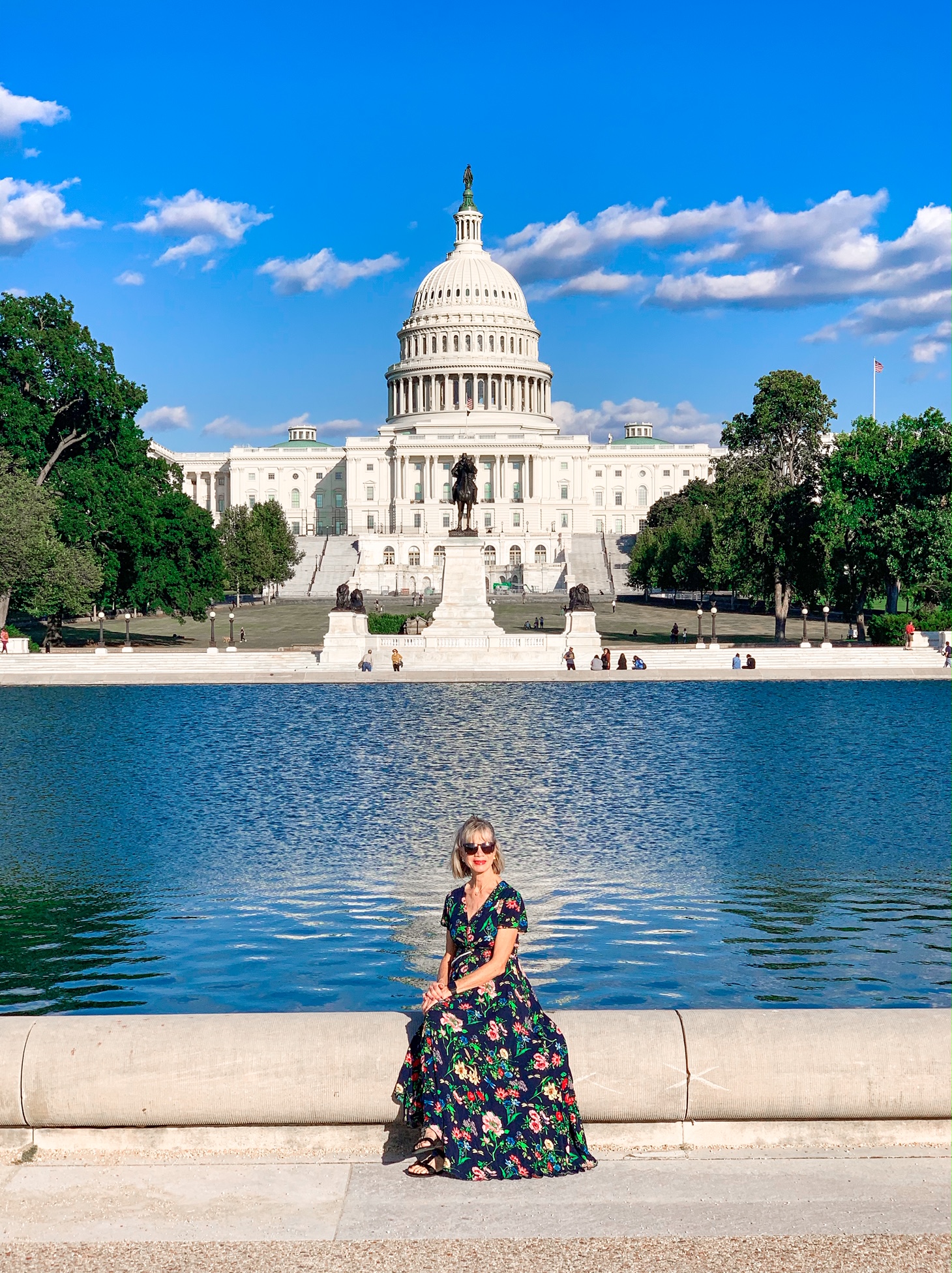 A woman in a dark floral dress sitting on the side of a reflecting pool in front of the United States Capitol Building. 