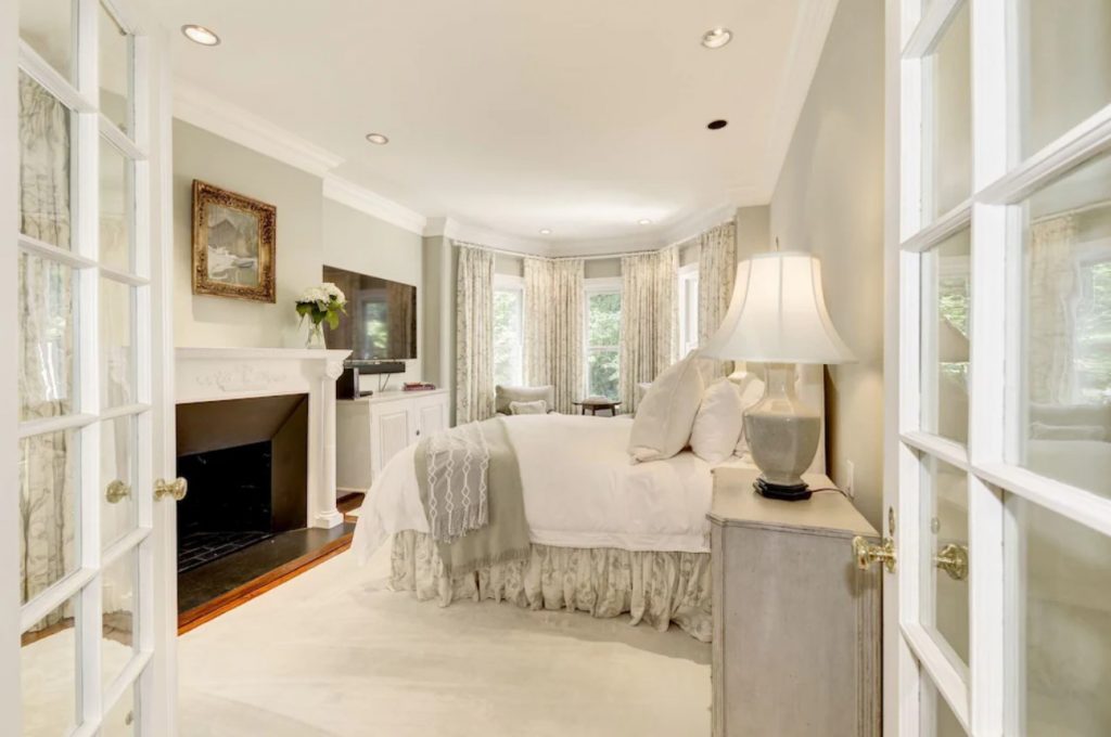 The bedroom of a VRBO in DC. It is mostly shades of white and cream with a gas-burning fireplace, large windows, and a large comfortable looking bed. 