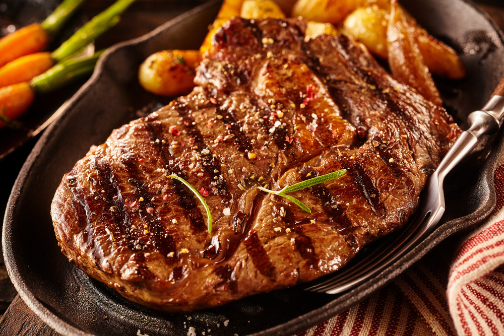 A grilled and seasoned rib eye steak in a cast iron pan, with vegetables in the background.