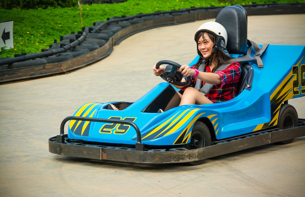 A woman in a blue go-kart driving down a paved road that is part of a go-karting course. 