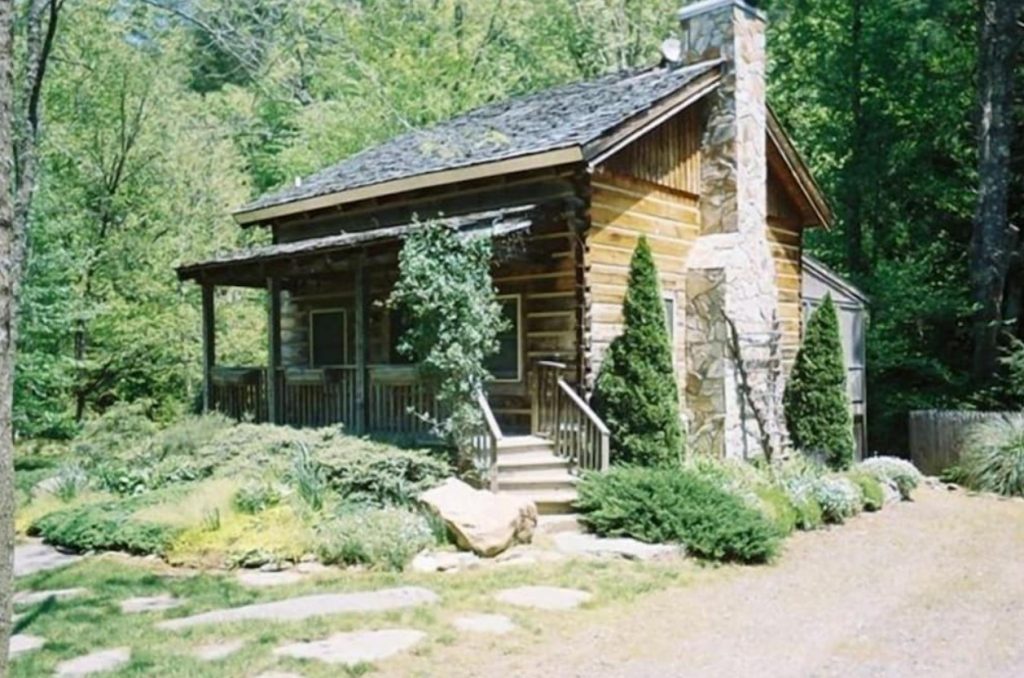 The exterior of an authentic log cabin. It has a large stone chimney, and is surrounded by a green garden and lots of trees. 