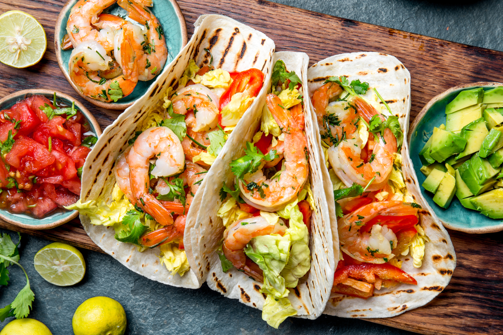 three shrimp tacos on wooden tray with bowls of sides surrounding the tacos