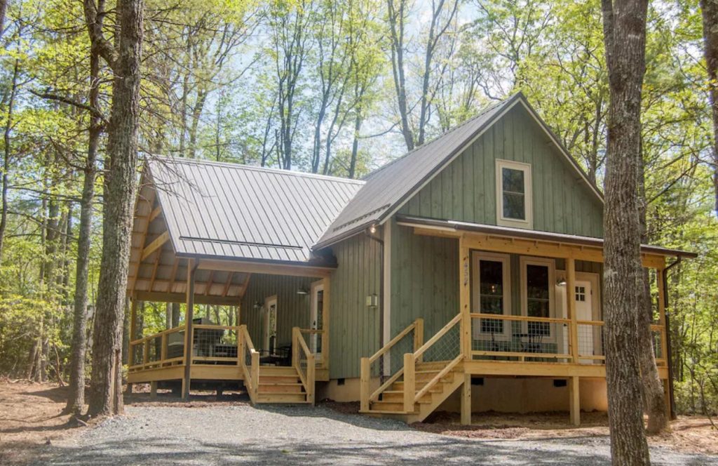The exterior of a brand new cabin in Boone. It is a light blue green and has a large side porch and a small front porch. Its surrounded by trees. 