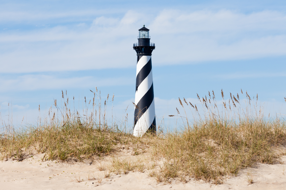 The iconic Cape Hatteras lighthouse behind some dunes. The dunes have tall grass growing on them. On the lighthouse are white and black stripes that wrap up the lighthouse. 