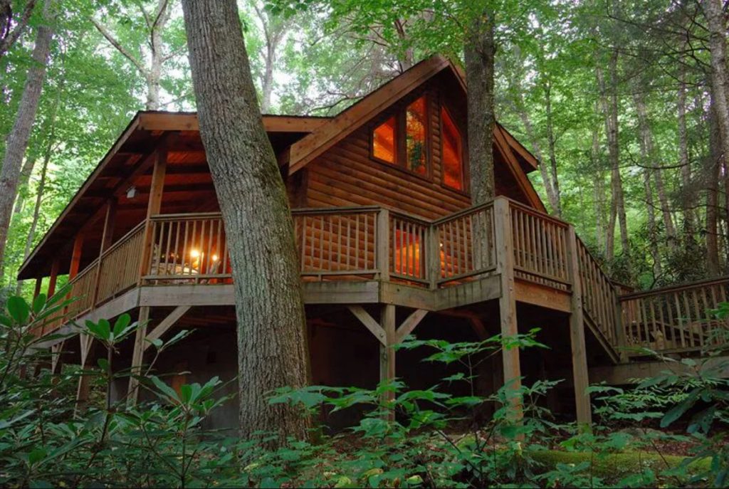 A classic log cabin nestled in the woods. It has a large wrap around deck and big windows in the peak of the roof.