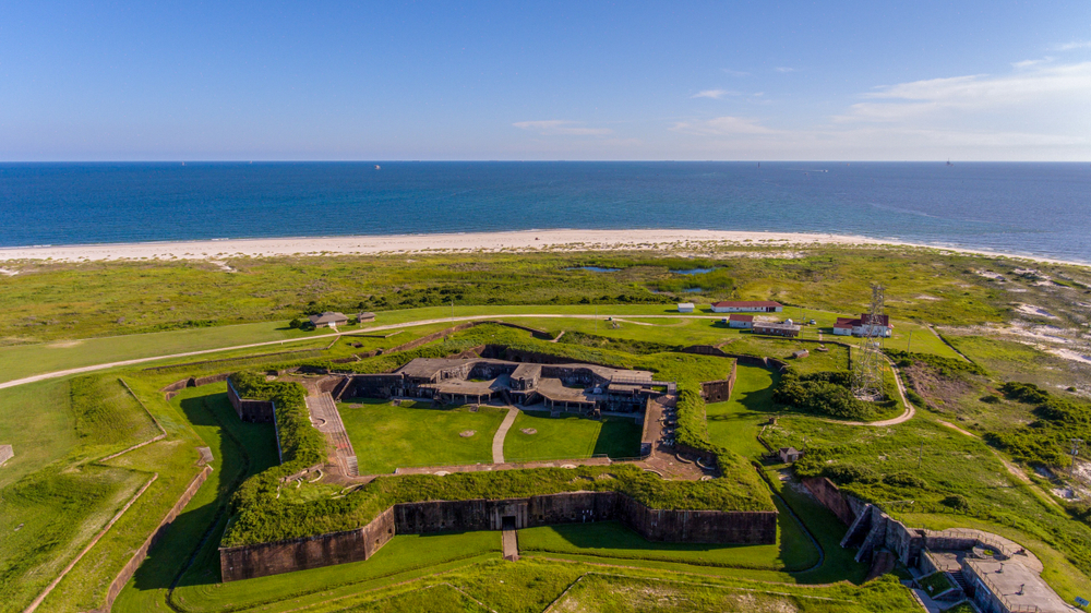 An aerial view of the old Fort Morgan that is covered with grass. Around it there is a manicured lawn. You can see a few other buildings and then the beach. 