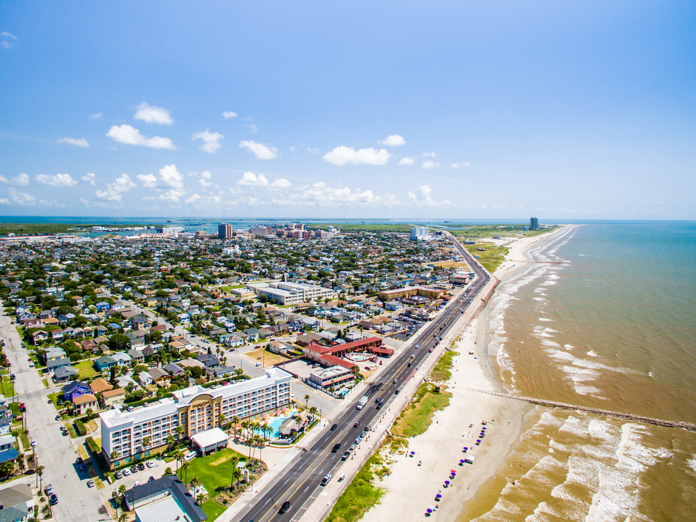 An aerial view of Galveston Texas. You can see the shoreline, lots of buildings and beach homes, and a long pier. Its one of the best islands in the south.