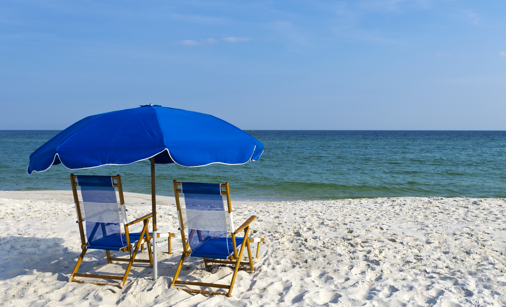 Two blue and white chairs under a blue beach umbrella. They are sitting on a white sand beach looking out at the Gulf of Mexico. 
