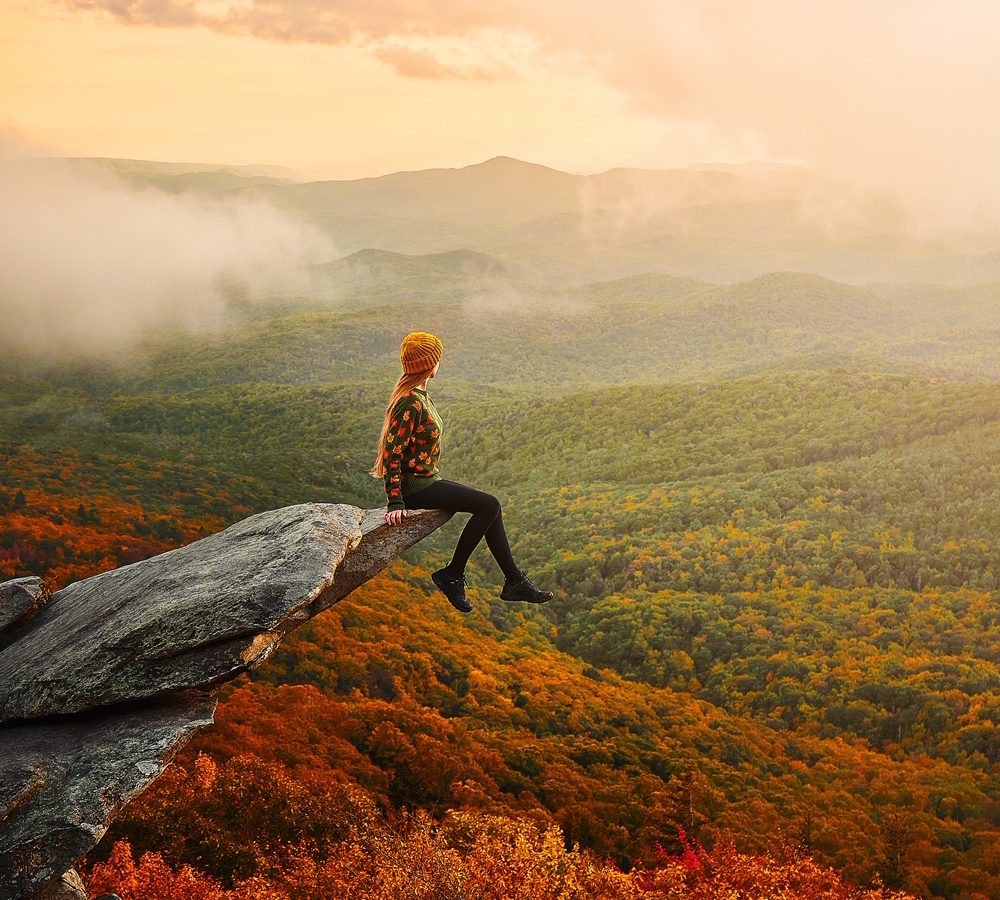 girl sitting on edge of large rock overlooking autumn valley while hiking in Boone