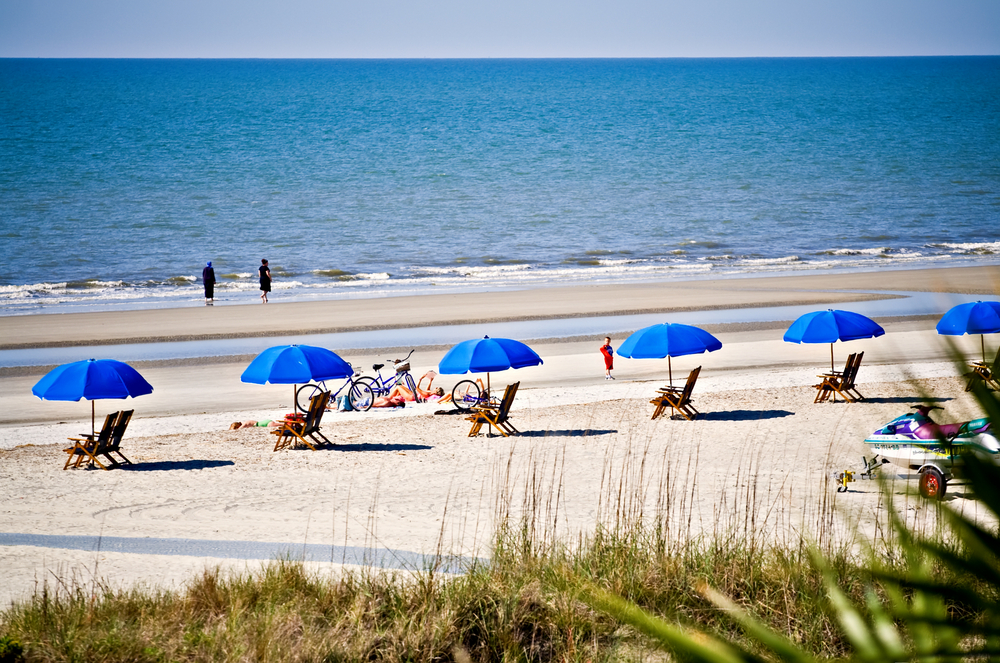 A row of beach chairs with blue umbrellas over them on a beach in Hilton Head South Carolina. There are also some bikes on the beach. You can see people walking on the shore as well. 