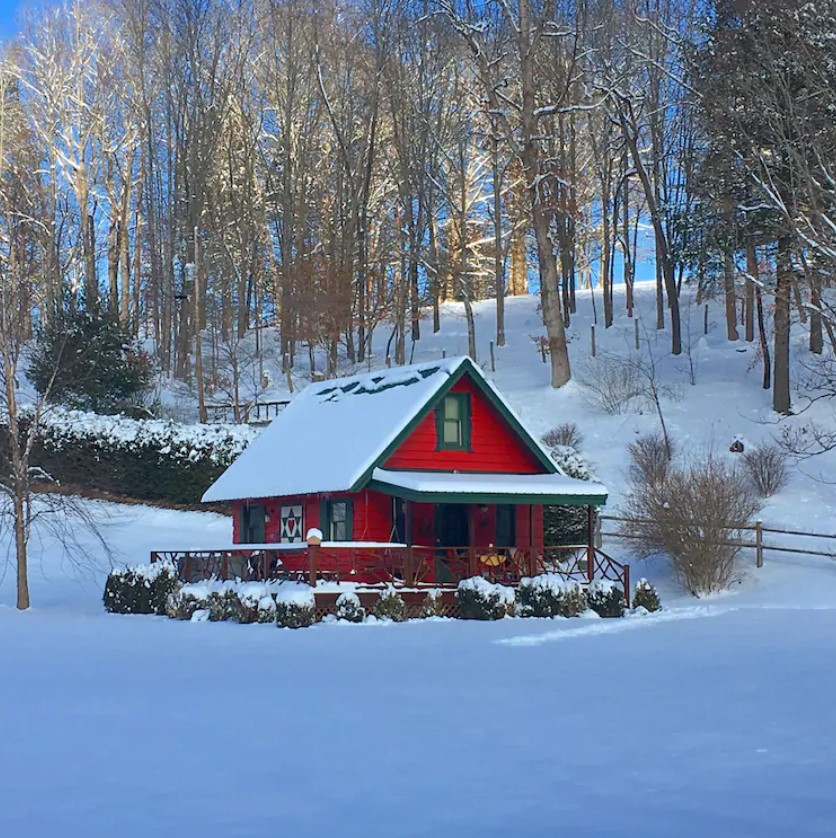 A small bright red cabins in Boone in the winter. There is snow covering the cabin, the land around it, and the trees on the hill behind it. 