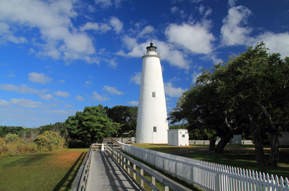 An old white lighthouse with a white picket fence around it. In the background you can see trees and shrubs. 