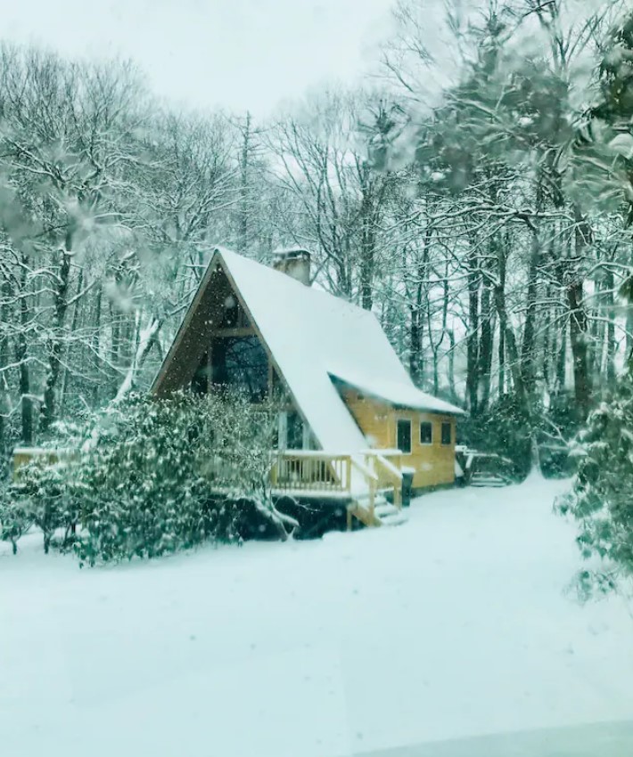 A classic A-frame cabin in Blowing Rock in the winter. The cabin, yard, and trees are covered in snow and it is kind of grey out. 