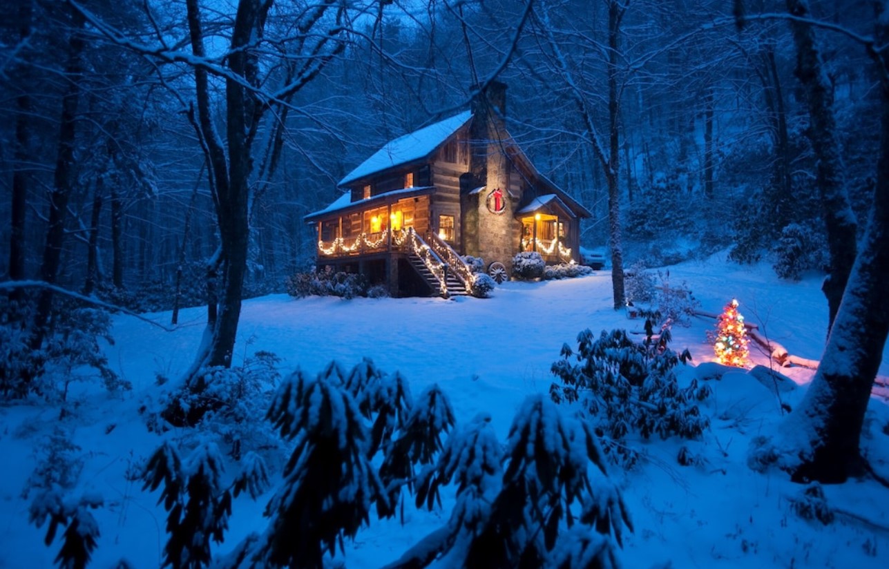 A log cabin in the woods at night in the winter. There is snow on the ground and the cabin has string lights on it. One of the best cabins in Boone.