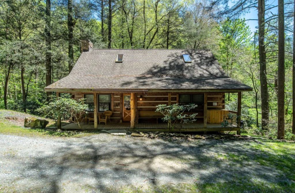 The front exterior of a classic log cabin. It has a small front deck with some chairs and a hot tub. Behind it you can see a dense forest. One of the best cabins in Boone. 