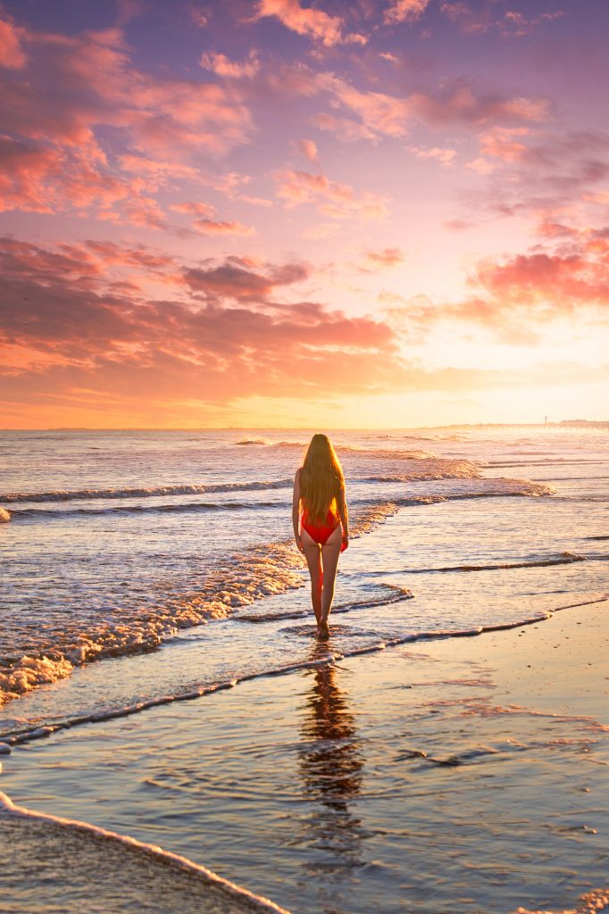 A woman in a red bathing suit walking down the shore of a beach at sunset. She is walking where the waves are gently coming up onto the shore. 