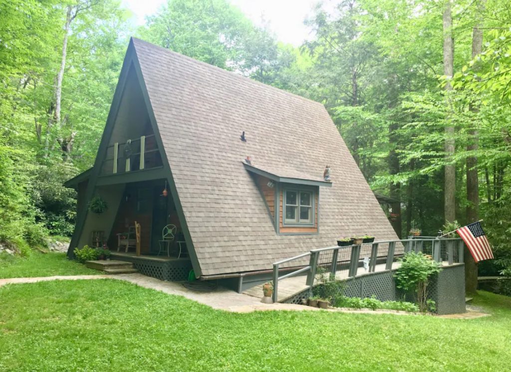 The exterior of a large A-frame cabin with a side porch, a front porch and deck, and a window on the side. It is surrounded by a grassy yard and trees. 