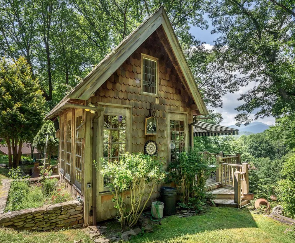 A small shingle cabin with lots of windows and a small porch and garden. You can see Grandfather Mountain in the distance behind it. 