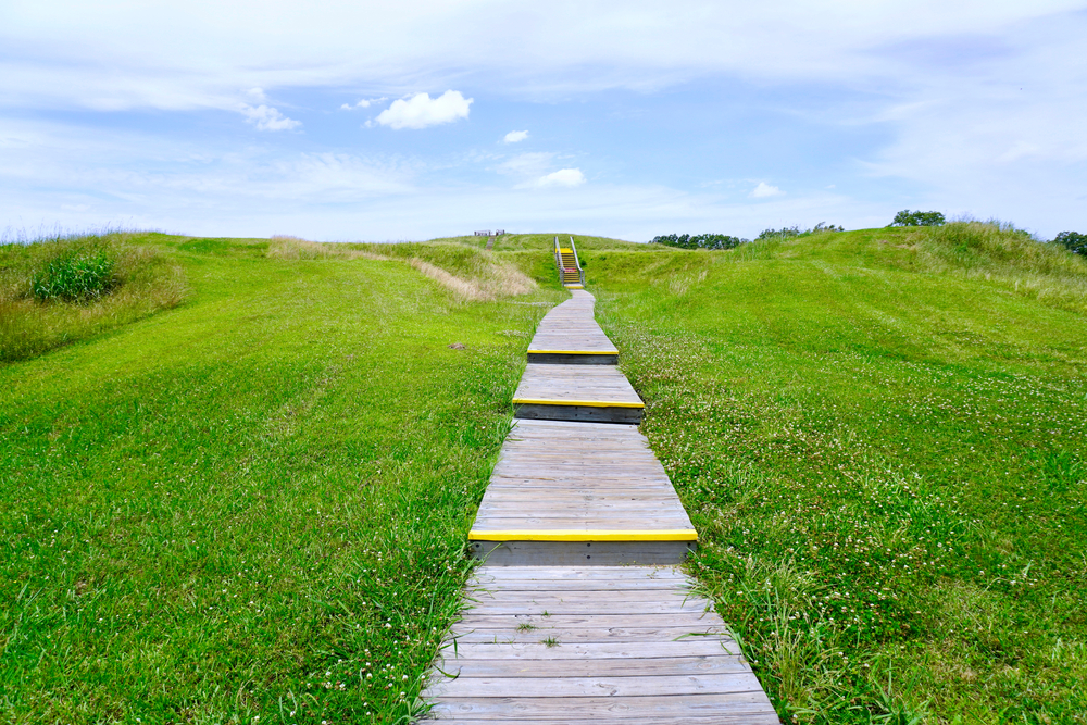 A wooden boardwalk sits atop one of the grass-covered earthen mounds at Poverty Point World Heritage Site, one of the best places to visit in Louisiana.