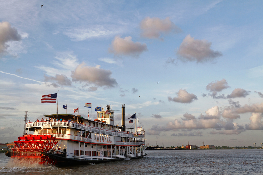 Steamboat Natchez, one of the best things to do in Louisiana, moves away from the camera along the Mississippi with flags and birds soaring in the breeze.