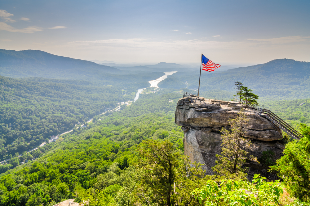 chimney rock, one of the best places to visit in North Carolina. American flag raised above the rock flowing in the wind with views of the mountains and river below