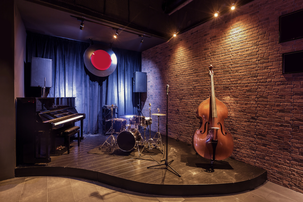The stage in a Jazz Club with a piano, drum set, and upright bass set up on it. There is a brick wall and what looks like a record hanging above a curtain in the back of the stage. 