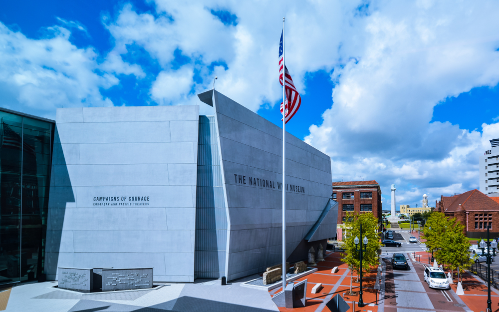 The exterior of the National WWII Museum in New Orleans. It is a large grey stone building with a red brick courtyard. There is an American Flag in front of the building. 