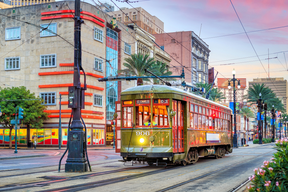 The St. Charles Streetcar on a street in New Orleans. The streetcar is green and red and is on a street surrounded by buildings and palm trees. 