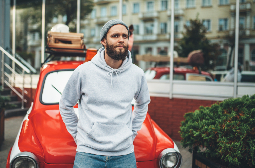 A man leans against a red car in a grey hoodie and Christmas trees are behind him, showing it's important to stay warm during winter in the south.