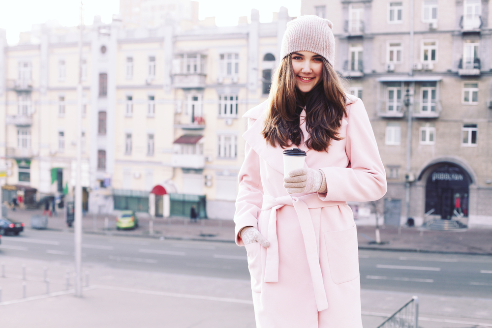 A woman holds a cup of coffee outside buildings in a warm, pink jacket, celebrating winter in the south.